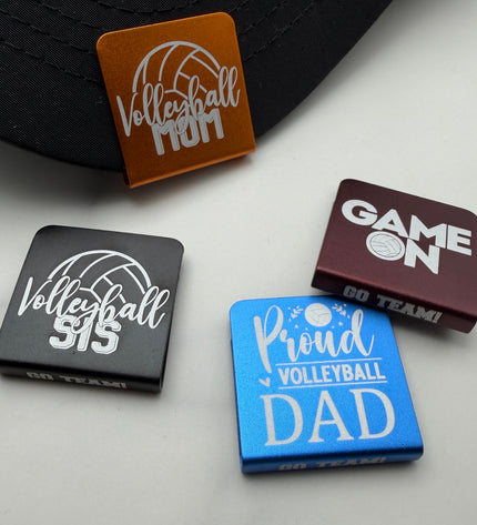 Four anodized aluminum hat clips with volleyball-themed designs are arranged on a light gray surface next to a black baseball cap. The badges are orange, black, maroon, and blue, each with white text and graphics reading phrases like 'Volleyball Mom,' 'Volleyball Sis,' 'GAME ON GO TEAM!,' and 'Proud VOLLEYBALL DAD.